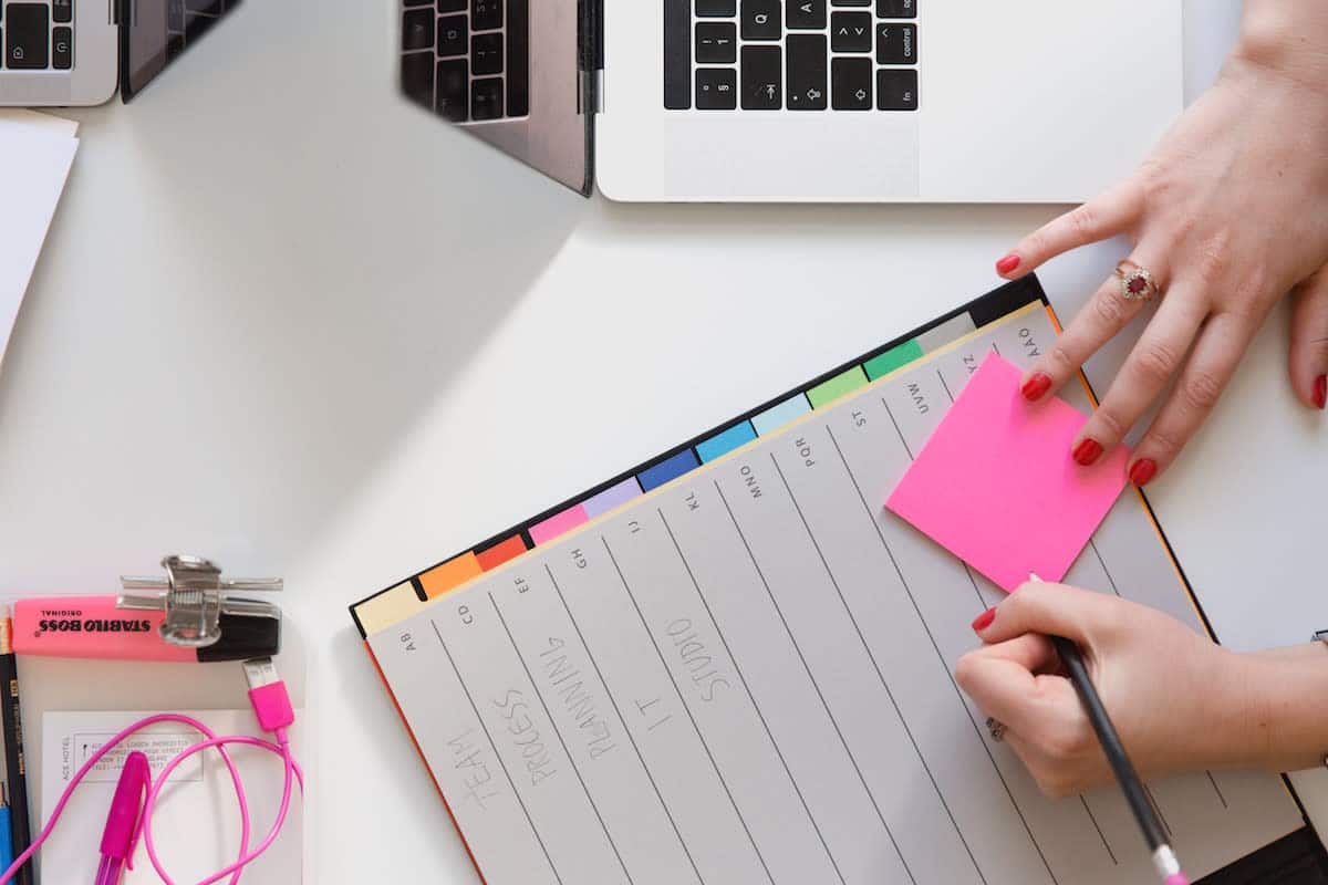 hands writing on a post-it note on a planner beside a laptop